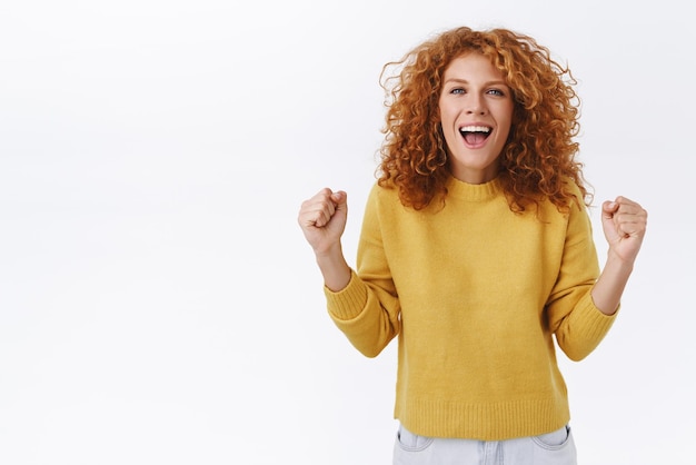 Animado alegre ruiva caucasiana encaracolada feminina na camisola amarela cerrar os punhos dançando em comemoração e alegria gritando palavras de torcida como sendo fã do time triunfando sobre a conquista do sucesso