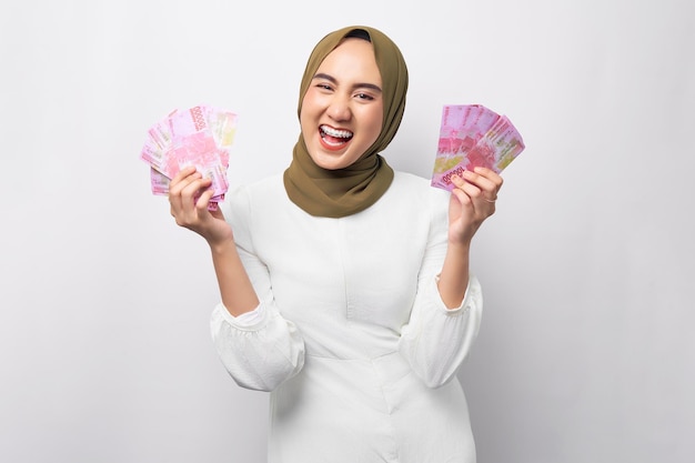 Animado bela jovem muçulmana asiática vestindo hijab segurando dinheiro em notas de rupia enquanto olha para a câmera isolada no fundo branco Conceito de estilo de vida religioso de pessoas