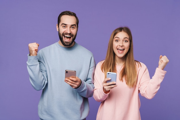 Foto animado casal amoroso surpreso isolado sobre a parede roxa usando telefones celulares faz gesto de vencedor.