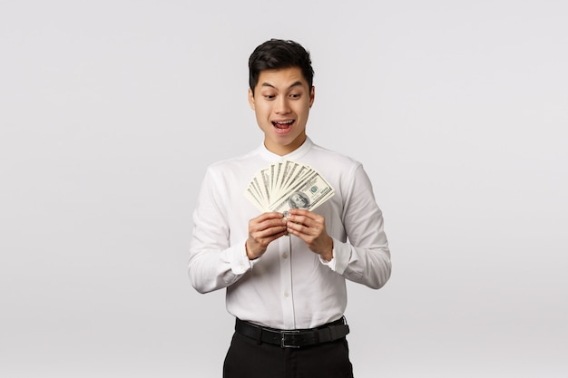 Animado e feliz, satisfeito, bonito homem de negócios asiático fez um bom negócio, contando dinheiro e sorrindo emocionado, ganhando lance, ganhou muito dinheiro e festejou, pronto para se tratar,