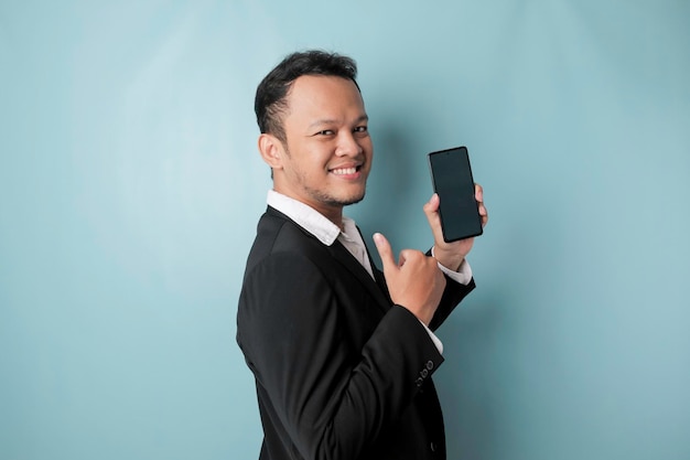 Animado empresário asiático vestindo terno preto dá polegares para cima gesto de aprovação enquanto mostra espaço de cópia em seu smartphone isolado por fundo azul