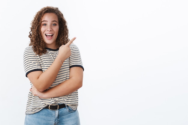 Foto animado, entusiasta, confiado, regordeta joven apuntando un enlace interesante que le muestra una promoción impresionante que indica la esquina superior derecha sonriendo emocionado, pared blanca