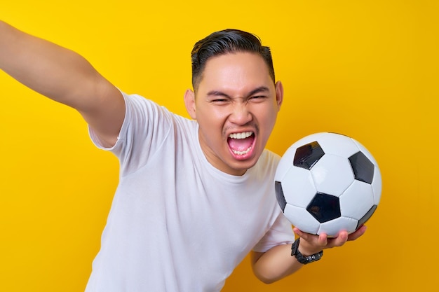 Animado fã de futebol jovem asiático em uma camiseta branca carregando uma bola de futebol e fazendo um tiro de selfie em um telefone celular isolado em fundo amarelo Conceito de estilo de vida de lazer de esporte de pessoas