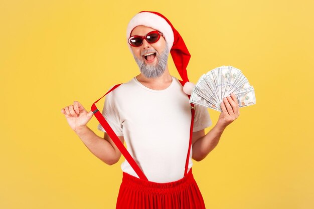 Animado homem idoso positivo com chapéu de papai noel e óculos de sol mostrando fã de dólares olhando para a câmera com expressão extremamente satisfeita, apostando. Tiro de estúdio interior isolado em fundo amarelo