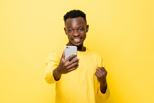 Animado jovem africano apertando o punho e olhando para o celular em fundo de estúdio amarelo isolado