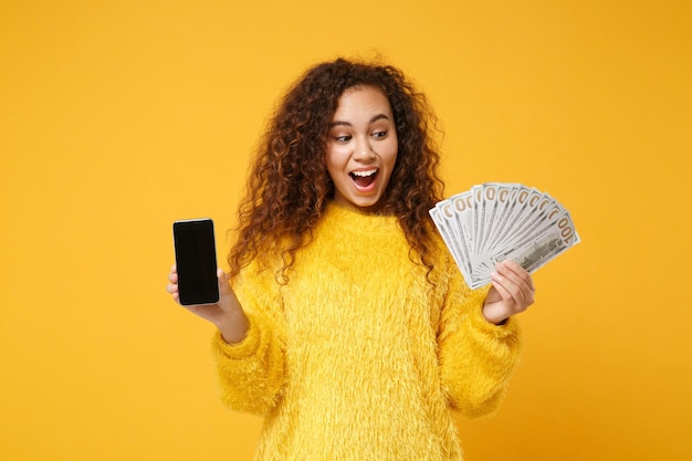 Animado jovem afro-americana posando isolado em fundo amarelo. Conceito de estilo de vida de pessoas. Mock up espaço de cópia. Segure o celular com o ventilador de tela vazia em branco de dinheiro em notas de dólar.