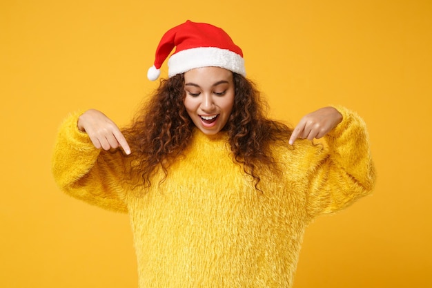 Animado jovem afro-americano Papai Noel em suéter de pele, chapéu de Natal isolado em fundo amarelo. Feliz ano novo 2020 conceito de feriado de celebração. Simule o espaço da cópia. Apontando os dedos indicadores para baixo.