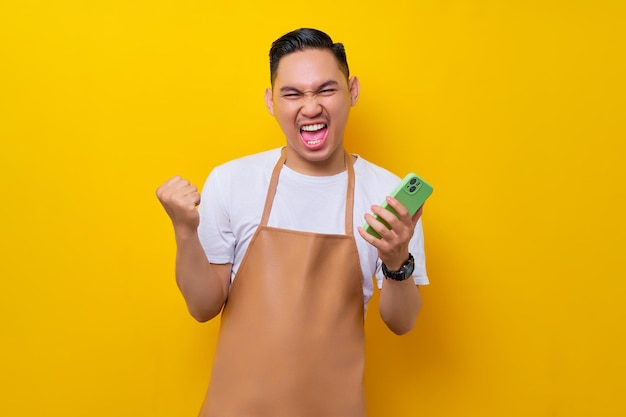 Animado jovem asiático barista barman empregado vestindo trabalho de avental marrom na cafeteria segurando o telefone móvel e mostrando o gesto vencedor isolado no fundo amarelo Conceito de inicialização de pequenas empresas