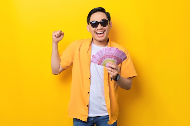 Animado jovem asiático bonito segurando notas de dinheiro e comemorando o sucesso isolado em fundo amarelo