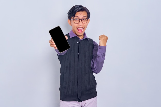 Animado jovem asiático em camisa casual e colete mostrando celular de tela em branco e fazendo gesto de vencedor isolado no fundo branco conceito de estilo de vida de pessoas