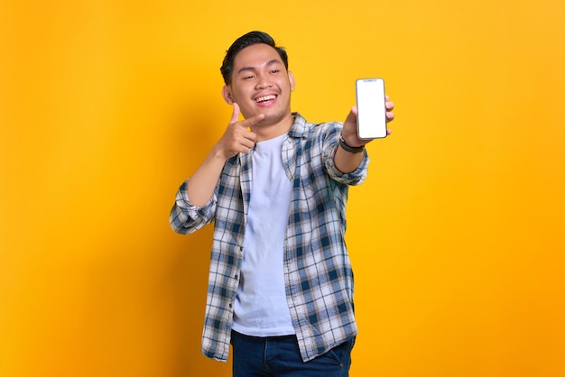 Animado jovem asiático em camisa xadrez mostrando tela em branco do celular recomendando aplicativo isolado em fundo amarelo