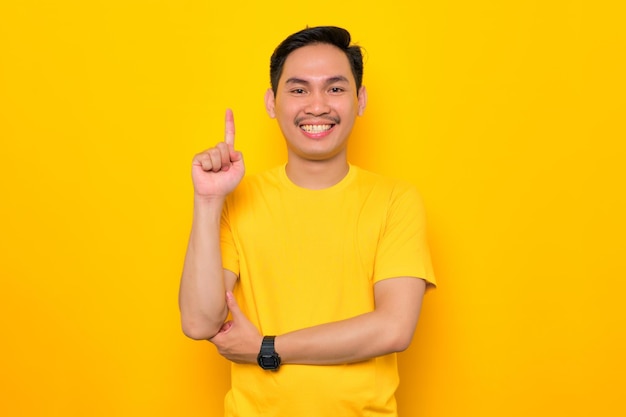 Animado jovem asiático em camiseta casual apontando o dedo para cima tendo uma ideia ou solução encontrada isolada em fundo amarelo Conceito de estilo de vida de pessoas