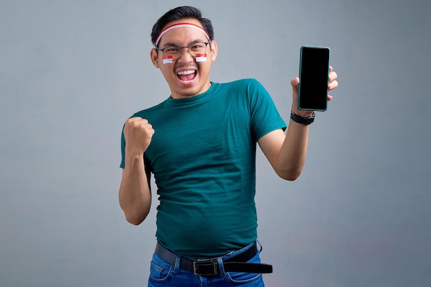 Animado jovem asiático em camiseta casual mostrando telefone celular com tela em branco, fazendo gesto de vencedor isolado no conceito de celebração do dia da independência indonésia de fundo cinza