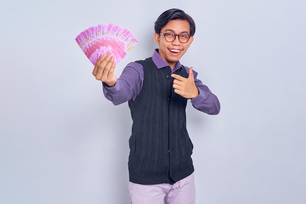 Animado jovem asiático em roupas de camisa roxa apontando dedos para dinheiro em notas de rupia isoladas em fundo branco Conceito de estilo de vida de pessoas