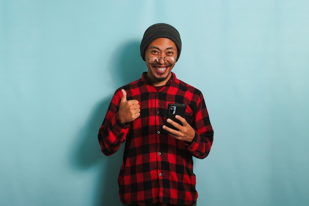 Animado jovem asiático está usando smartphone e mostrando um polegar para cima sinal isolado no fundo azul