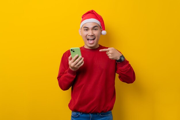 Animado jovem asiático no chapéu de Papai Noel apontando o dedo para smartphone recomendando aplicativo móvel na celebração de fundo amarelo estúdio feriado de Natal e conceito de ano novo