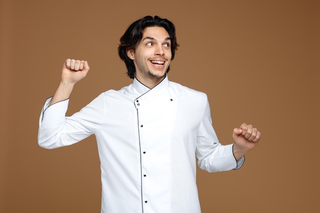 animado jovem chef masculino vestindo uniforme olhando para o lado mantendo os punhos no ar isolados no fundo marrom