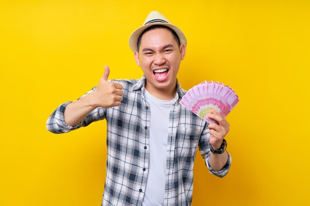 Animado jovem de etnia asiática 20 anos vestindo roupas casuais chapéu segurando na mão leque de dinheiro em notas de rupia mostra polegar para cima gesto isolado em fundo amarelo Pessoas conceito de estilo de vida