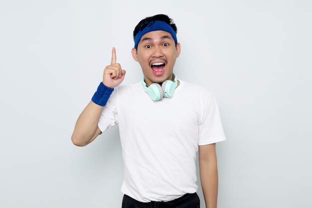 Animado jovem desportista asiático na faixa azul e camiseta branca de roupas esportivas com fones de ouvido apontando os dedos para cima tendo uma boa ideia isolada no fundo branco conceito de esporte de treino