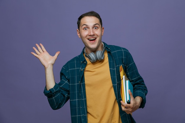 Animado jovem estudante do sexo masculino usando fones de ouvido no pescoço, segurando as ferramentas de estudo debaixo do braço, olhando para a câmera mostrando a mão vazia isolada no fundo roxo