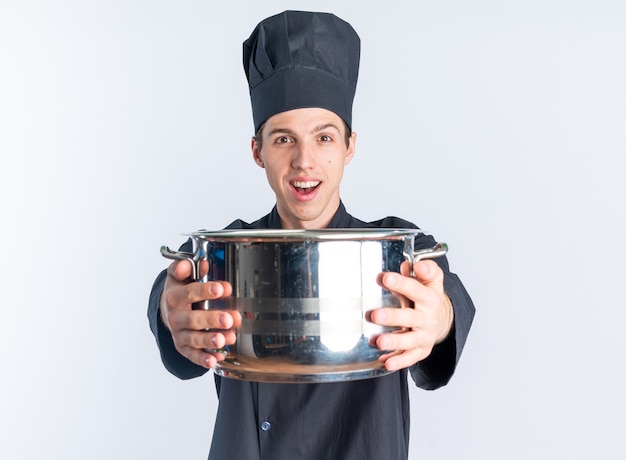 Animado jovem loiro cozinheiro masculino com uniforme de chef e boné estendendo a panela em direção à câmera, olhando para a câmera isolada na parede branca