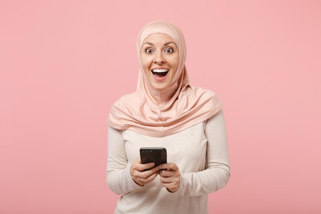 Animado jovem muçulmana árabe em roupas leves hijab posando isolado no fundo rosa. Conceito de estilo de vida do Islã religioso de pessoas. Mock up espaço de cópia. Usando o telefone celular, digitando mensagem sms.