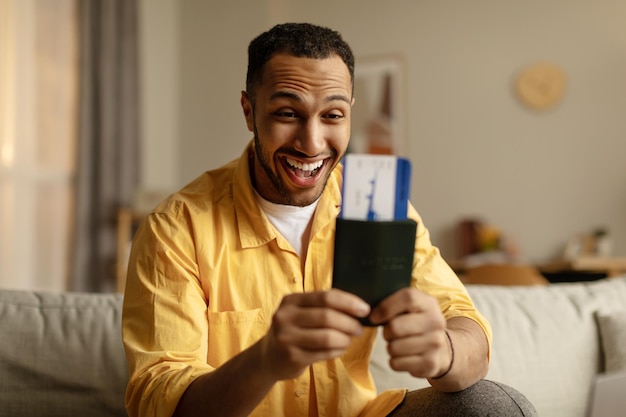 Animado jovem negro com passaporte e bilhetes se preparando para as férias de verão sentado no sofá