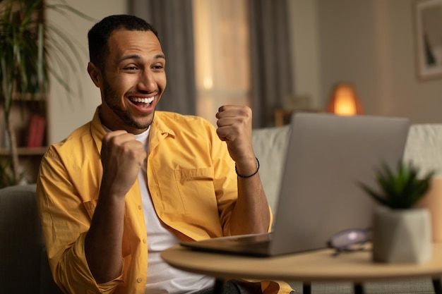 Animado jovem negro olhando para a tela do laptop gesticulando SIM, sentindo-se feliz com a vitória ou promoção on-line em casa