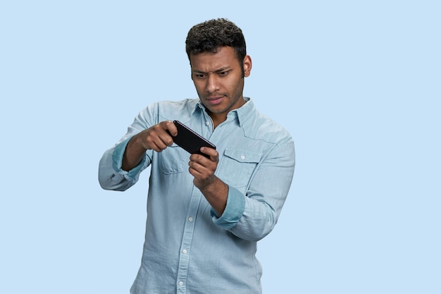 Animado jovem sul-asiático jogando jogo em videogame móvel isolado em fundo azul pastel