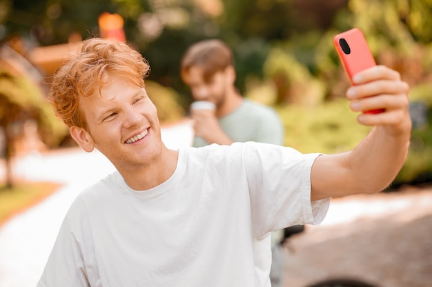 Foto animado lindo jengibre joven caucásico fotografiando a sí mismo con un teléfono inteligente con su amigo en el fondo