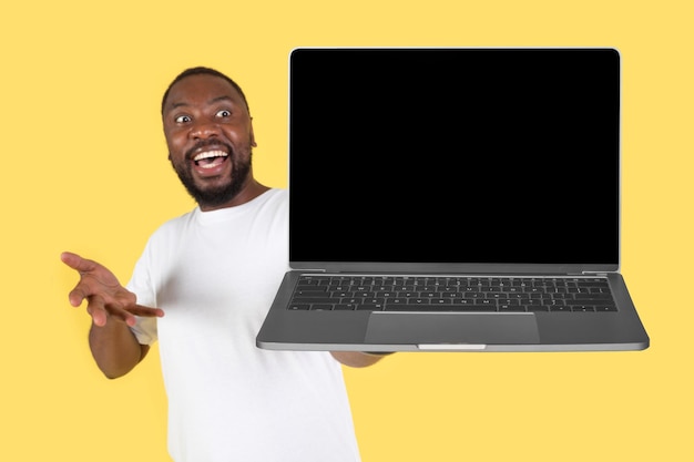 Animado macho afro-americano mostrando computador portátil sobre fundo amarelo