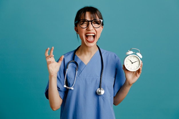 Animado segurando despertador jovem médica usando estetoscópio uniforme de quinto isolado em fundo azul