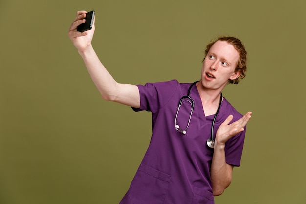 Animado, tire uma selfie jovem médico masculino vestindo uniforme com estetoscópio isolado em fundo verde