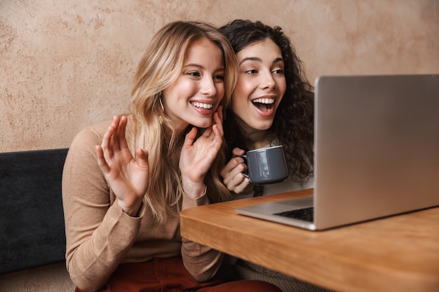 animados amigos de meninas bonitas felizes sentados em um café usando um laptop acenando e conversando