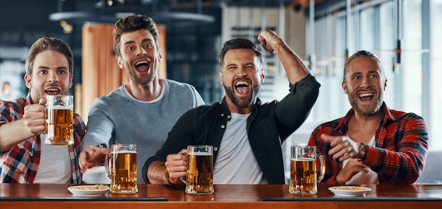 Animando a los hombres jóvenes en ropa casual bebiendo cerveza y viendo juegos deportivos