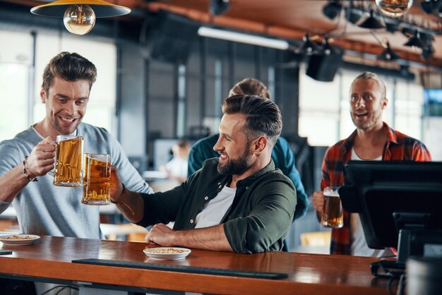 Animando a los hombres jóvenes en ropa casual viendo juegos deportivos y disfrutando de la cerveza mientras están sentados en el pub