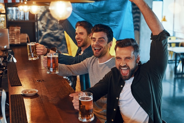 Animando a los jóvenes cubiertos con la bandera de Ucrania bebiendo cerveza y viendo un juego deportivo mientras están sentados en el pub