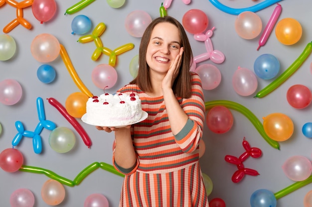 Aniversariante alegre Mulher sorridente satisfeita com cabelo castanho, vestido listrado, olhando para a câmera com cara feliz, segurando o bolo em pé contra a parede cinza decorada com balões coloridos