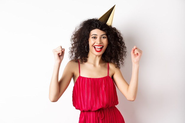 Aniversariante com chapéu de festa se divertindo, dançando com um vestido vermelho e cantando, em pé contra um fundo branco.