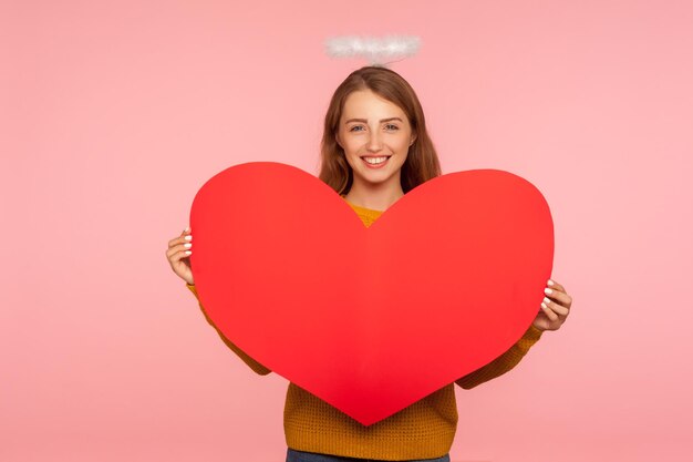 Anjo no dia dos namorados39s Retrato de uma linda garota ruiva com auréola na cabeça sorrindo para a câmera e segurando o grande símbolo do coração vermelho de amor e carinho sentimentos românticos studio shot fundo rosa