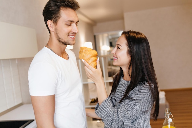 Anmutige junge Frau in grauer Kleidung, die morgens Freund mit Brötchen füttert. Romantisches dunkelhaariges Mädchen mit aufrichtigem Lächeln, das Croissants mit Ehemann genießt.