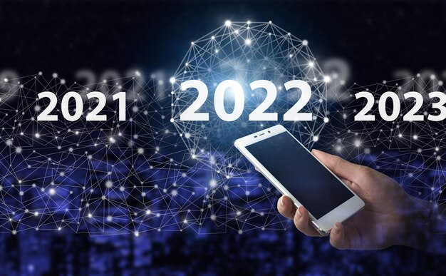 Año de carga 2021 a 2022. Concepto de inicio. Asimiento de la mano smartphone blanco con señal de holograma digital 2022 en la ciudad de fondo borroso oscuro. Concepto de visión 2021-2022.