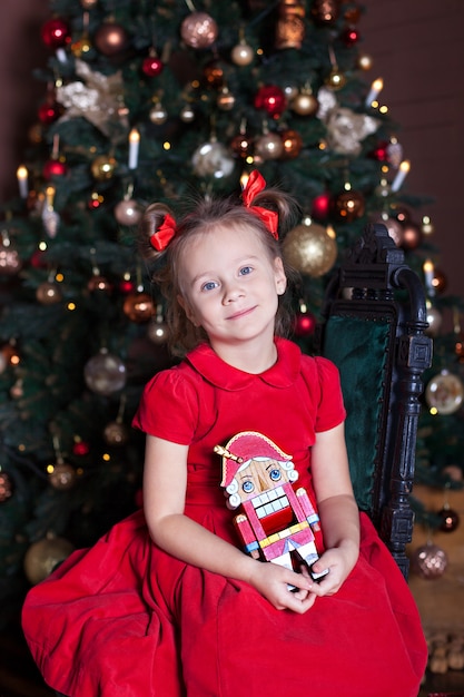 Ano Novo 2020! Feliz Natal, boas festas! Uma menina adorável com um quebra-nozes nas mãos se senta em uma cadeira no interior de um ano novo lindamente decorado com uma árvore de Natal. Inverno