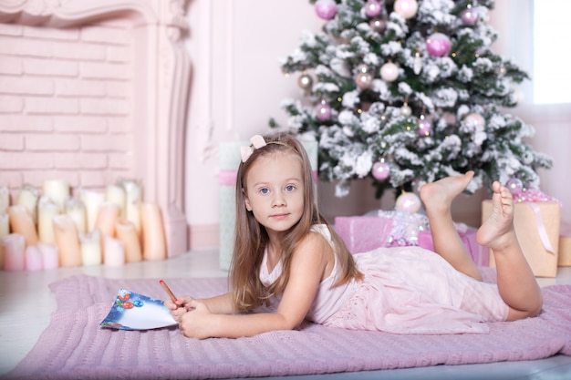 Ano novo 2020! o conceito de natal, férias e infância. menina bonita escreve uma carta para o papai noel perto da árvore de natal no quarto das crianças. menina esperando o natal em casa