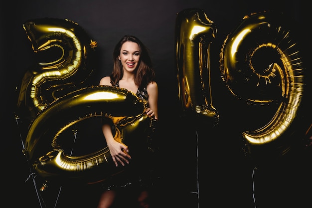 Ano novo. mulher com balões comemorando na festa. retrato da bela menina sorridente no vestido brilhante, jogando confetes, se divertindo com balões de ouro 2019 no fundo. alta resolução.