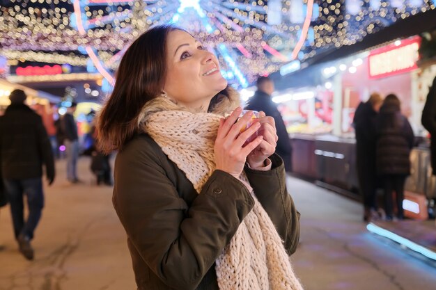 Ano Novo, Natal, mercado noturno festivo da cidade, mulher madura e feliz com uma caneca de vinho quente