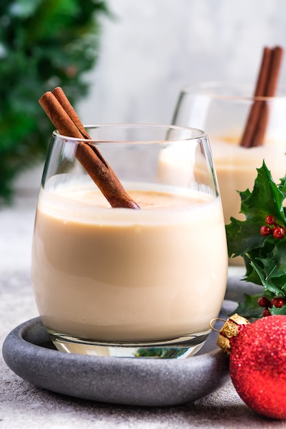 Ano novo ou coquetel de gemada de Natal com canela e noz-moscada em um copo, galhos de bagas de azevinho e bola brinquedo na pedra clara, decoração festiva