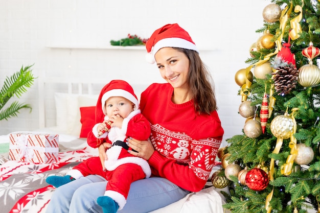 Ano Novo ou Natal, uma jovem mãe com um bebê na cama em casa perto da árvore de Natal disfarçada de Papai Noel abraçando e esperando o feriado sorrindo