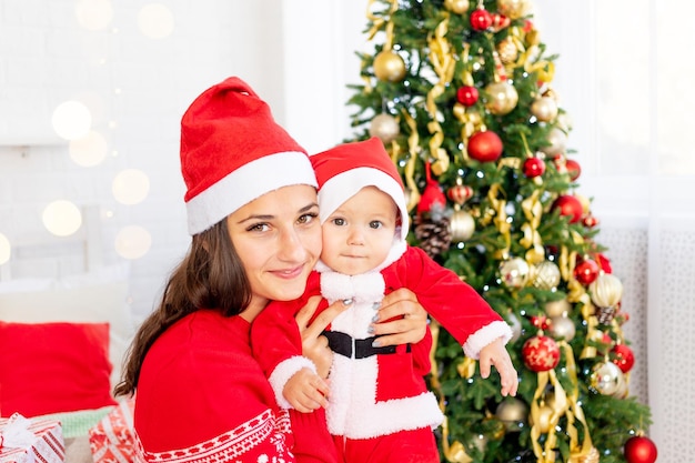 Ano novo ou natal, uma jovem mãe com um bebê na cama em casa perto da árvore de natal fantasiada de papai noel abraçando e esperando o feriado sorrindo