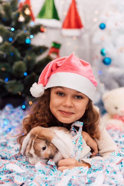 Año nuevo, una niña con un gorro de Papá Noel rosa se encuentra cerca de un árbol de Navidad y acaricia un conejo